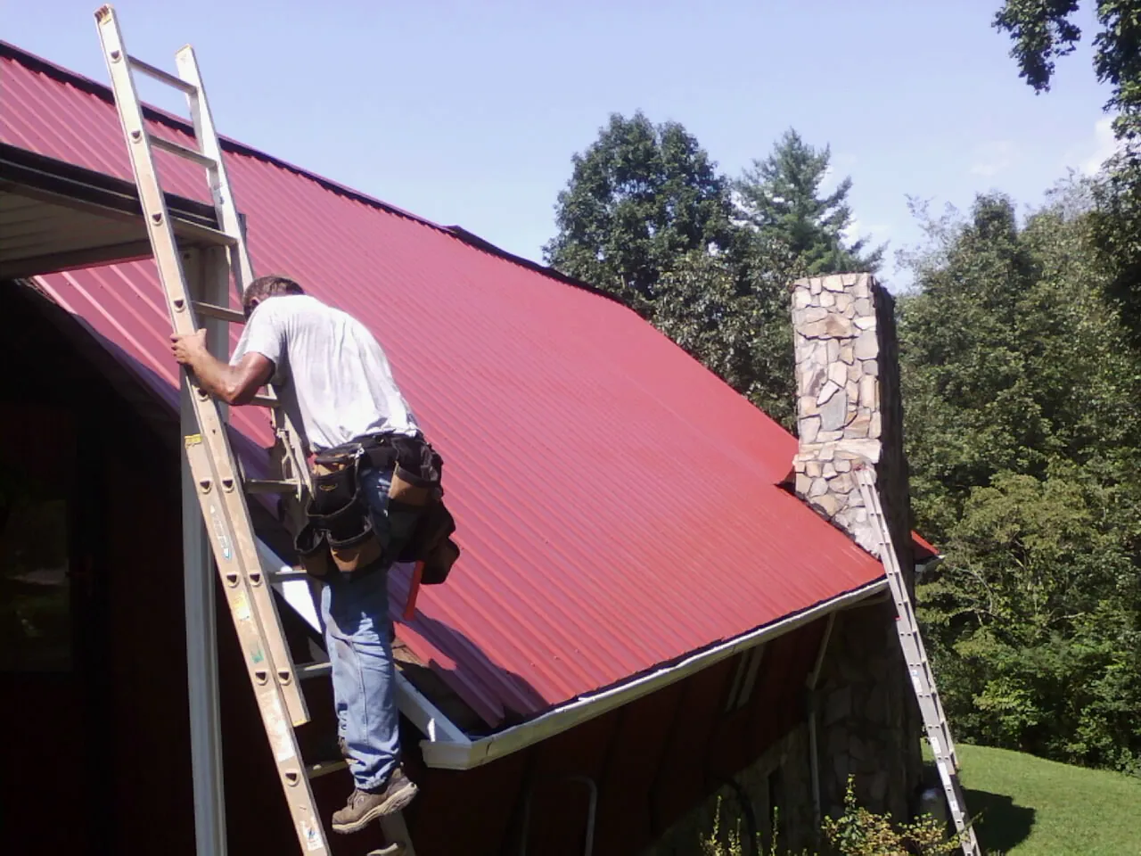 Roofer on a ladder beside a red metal roof and stone chimney — Soffit & Fascia Repair by Roofing Brothers in Olivet